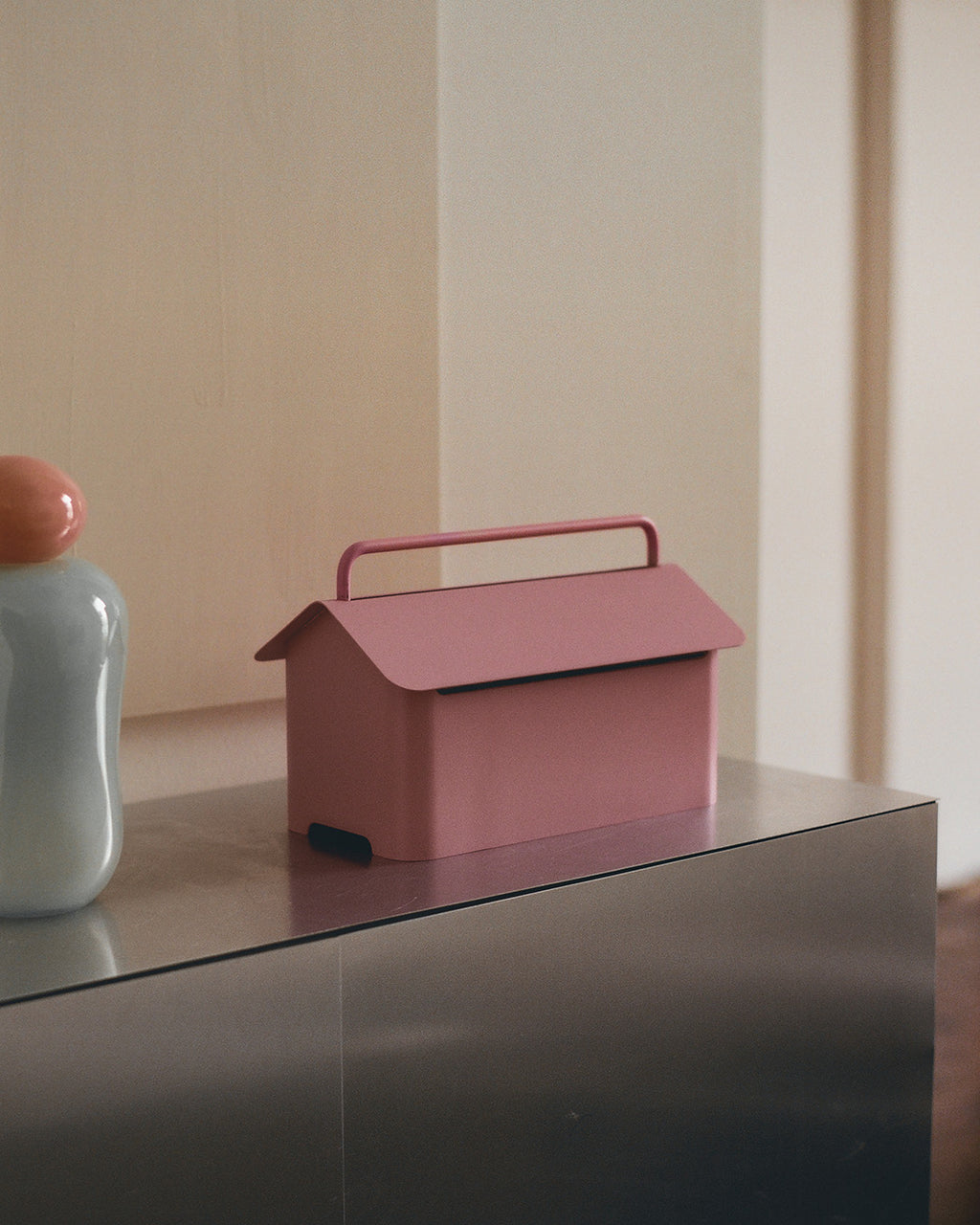 Cable Villa in Bubble Gum op een metalen dressoir naast een vaas, huisvormig kabelbeheer met handgreep in zijaanzicht